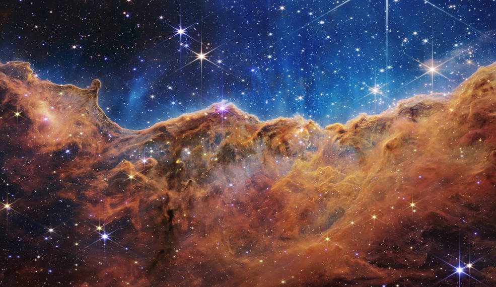 An undulating, transparent star-forming region of the Carina Nebula is shown in this web image, colored in blue and blue skies.  Foreground stars with diffraction heights can be seen, and patches of light in the background can be seen through the cloudy nebula.