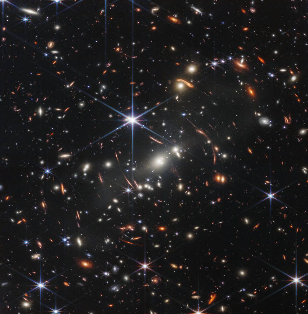 Distant galaxies appear as bright, glowing spots in this Webb telescope image, some smudged by a gravitational lens;  Foreground stars appear bright with hexagonal diffraction spikes, due to the shape of Webb's mirrors