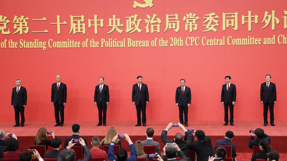 Seven Chinese politicians stand on stage in front of journalists, who photograph them with their mobile phones. 
